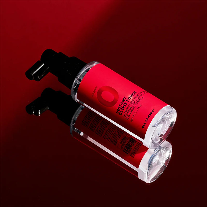 Flacone trasparente con etichetta rossa con scritte nere e tappo spray nero su tavolo in vetro che riflette un telo rosso.